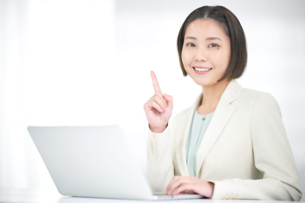 ノートパソコンを前に人差し指を立てる白いジャケットの女性