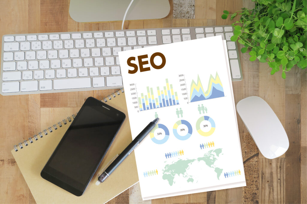 ブログSEOの資料とパソコンとスマホとノートとペンと植物-茶色い木目デスク